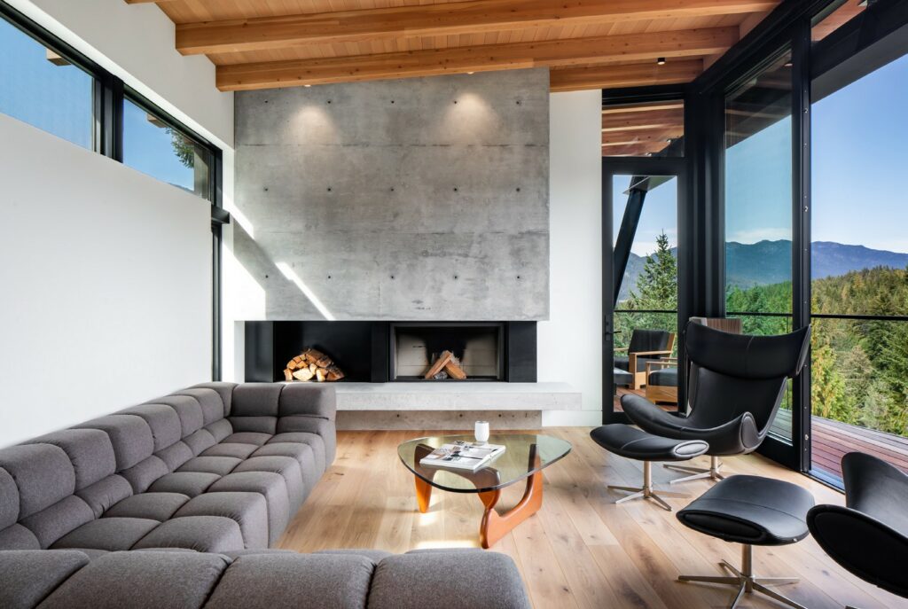 Modern living room designed by Custom Building Network with a concrete fireplace, gray sectional sofa, glass coffee table, black lounge chairs, wood ceiling, and large windows overlooking trees and mountains.