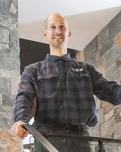 A man wearing a plaid shirt stands on a staircase in a building with stone walls, looking at the camera and smiling.