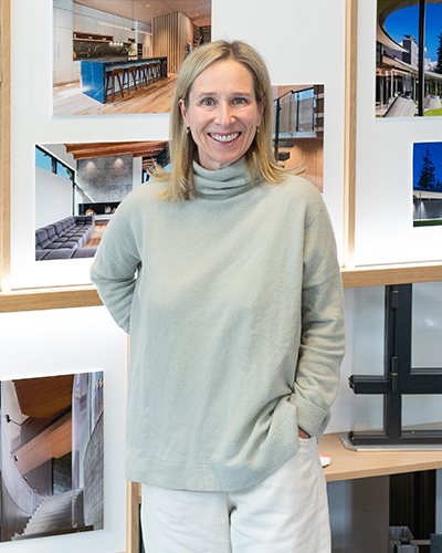 A woman in a light gray turtleneck sweater stands smiling in front of a wall displaying architectural photographs.