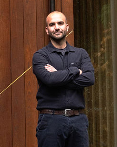 A man with a shaved head stands in front of a wooden wall with his arms crossed, wearing a dark long-sleeve shirt and pants.