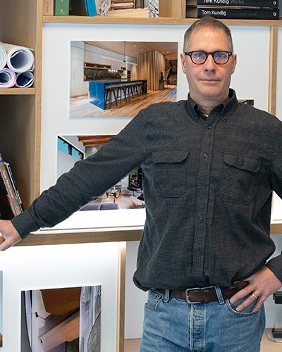 A man in glasses and a dark shirt stands in front of a wall displaying architectural photos and shelves with books and rolled blueprints.