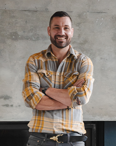 A man with a beard and short hair stands smiling with arms crossed, wearing a grey and yellow plaid shirt in front of a concrete wall.