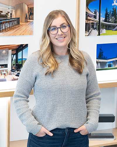 A woman with long, wavy blonde hair and glasses stands indoors, wearing a gray sweater and jeans, with architectural photos displayed on the wall behind her.
