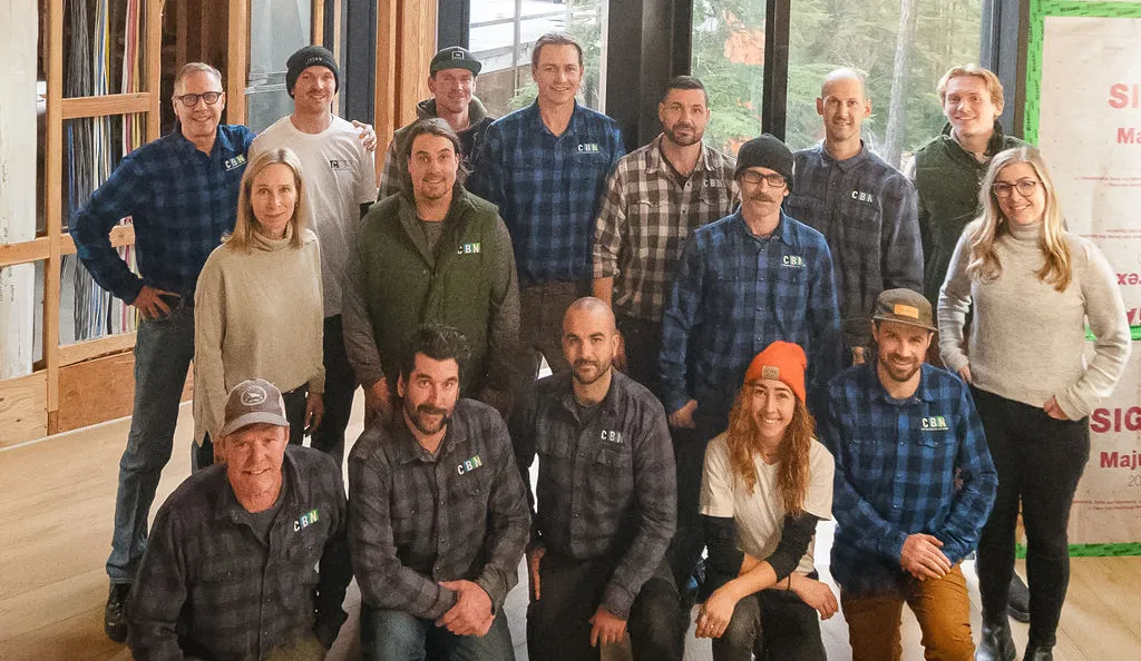 A group of sixteen adults pose indoors at a Custom Building Network event, with some standing and others kneeling, dressed in casual clothing including plaid shirts, hats, and beanies.