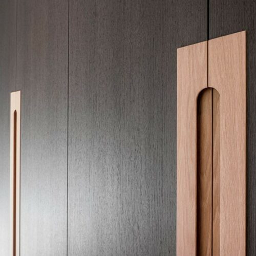 Close-up of modern wooden cabinet doors with vertical recessed handles, set against a dark wood finish, with a window and greenery visible on the left.