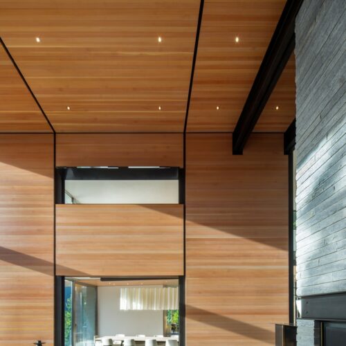 Modern interior with high wood-paneled ceilings, large windows, gray stone fireplace, and a dining area visible through an open passageway.