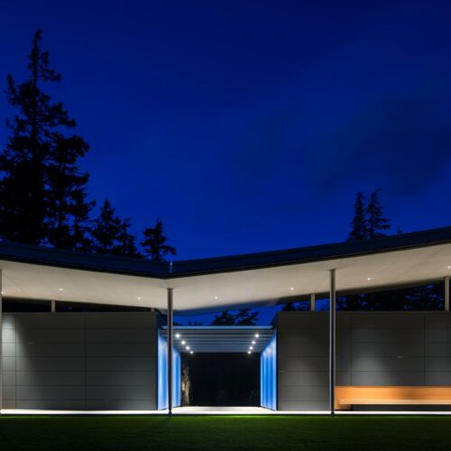 Modern building exterior at night with illuminated overhangs, sleek gray walls, and an open courtyard; tall trees are visible in the background under a deep blue sky.