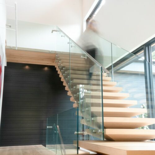 A person ascends a modern floating wooden staircase with glass railings in a bright, contemporary interior space.