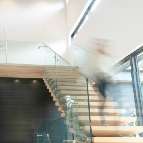 A person descends a modern wooden staircase with glass railings in a bright, spacious interior with large windows and exposed wooden ceiling beams.