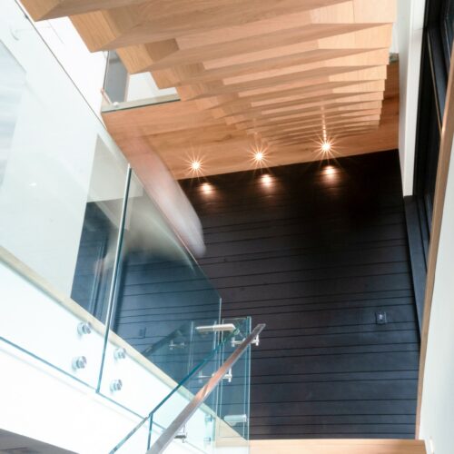 Modern wooden staircase with glass railing, viewed from below, featuring recessed wall lights and dark vertical paneling on the wall.