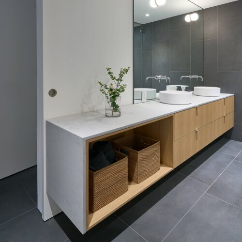 Modern bathroom with a long wood vanity, dual vessel sinks, large mirror, gray tile flooring, and two wicker storage baskets on an open shelf.
