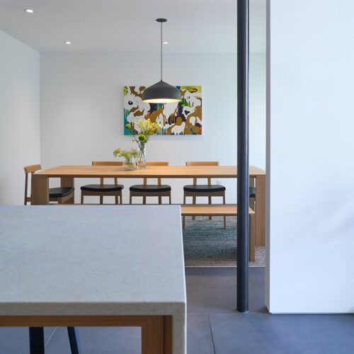 Modern dining room with a wooden table, six chairs, pendant light, wall art, and a vase of flowers, viewed from a kitchen counter with a black stool.