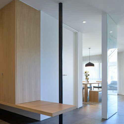 Modern interior hallway with floating wooden bench, vertical black metal support, tiled floor, and view into a dining area with a table, chairs, and pendant light.