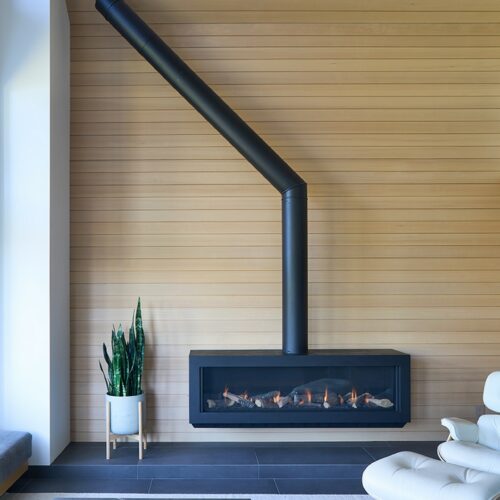Modern living room with a black wall-mounted fireplace, angled chimney pipe, light wood paneling, potted plants, and a white lounge chair.
