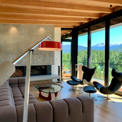 Modern living room with a gray sofa, floor lamp, black lounge chairs, fireplace, large windows, wooden ceiling, and mountain view in the background.