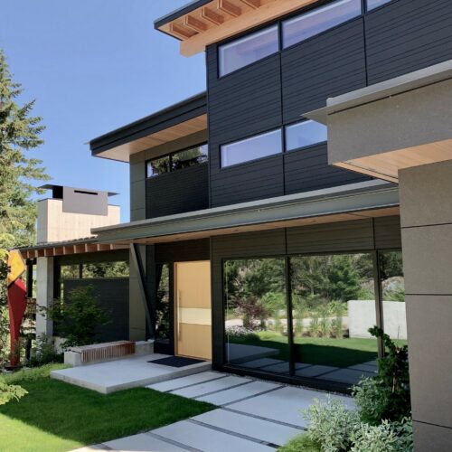 Modern two-story house with large windows, dark horizontal siding, light wood accents, and a landscaped front yard under a clear sky.