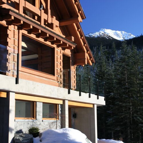 A wooden and concrete chalet with glass railings sits in a snowy, forested mountain area, with a snow-covered peak visible in the background under a clear blue sky.