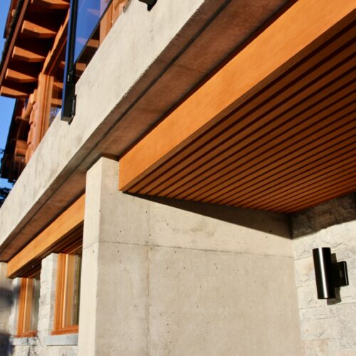 Close-up of a modern house exterior featuring concrete walls, wood trim, a wooden slatted ceiling, and large windows under bright sunlight.