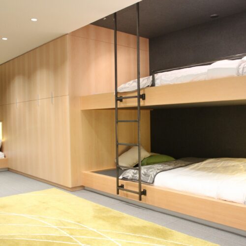 Modern bedroom with built-in wooden bunk beds and a single bed, featuring a yellow rug, gray walls, and recessed lighting.