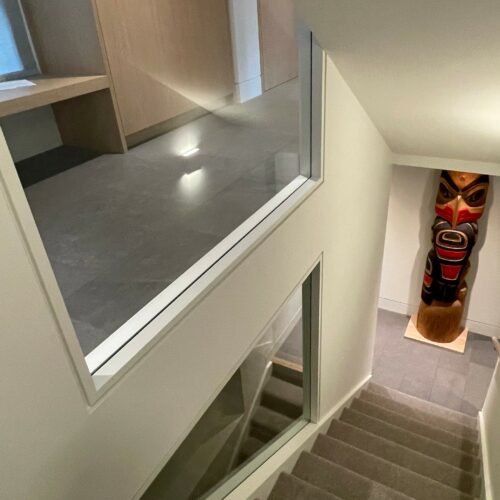 A view down a carpeted staircase with two interior windows; at the bottom of the stairs is a decorative totem pole on a tiled floor.