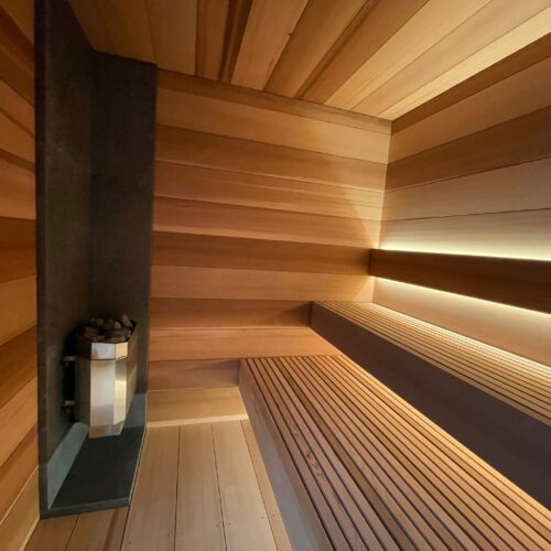 A modern wooden sauna with tiered bench seating, indirect lighting, and a metal heater with stones in the corner.