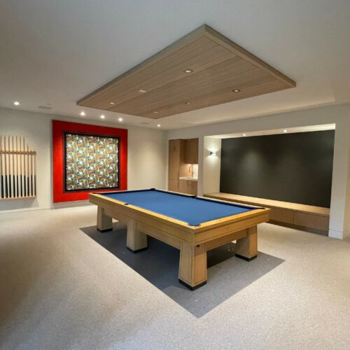 A spacious room with a pool table at the center, wall-mounted pool cues, a colorful wall art, a bench, and carpeted flooring.