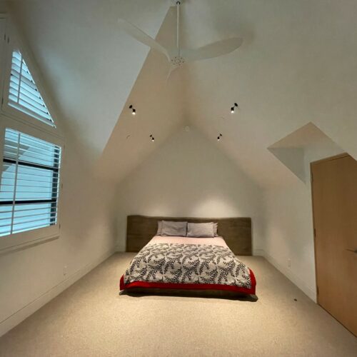 A minimalist bedroom with a bed centered against the back wall, a wooden door on the right, and slanted ceilings with a ceiling fan and recessed lighting.