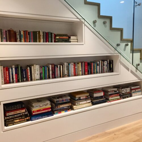 Built-in bookshelves under a modern staircase display an array of books and magazines arranged neatly on three staggered shelves.
