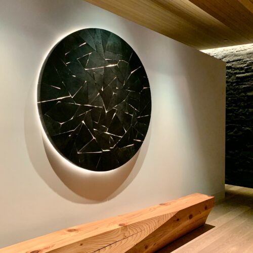 A circular black abstract wall sculpture with geometric patterns hangs above a wooden bench in a modern, minimalist hallway.