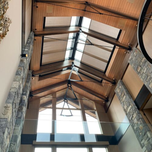 Wood and stone interior with a high vaulted ceiling, exposed beams, large windows, and a glass railing, viewed from below.