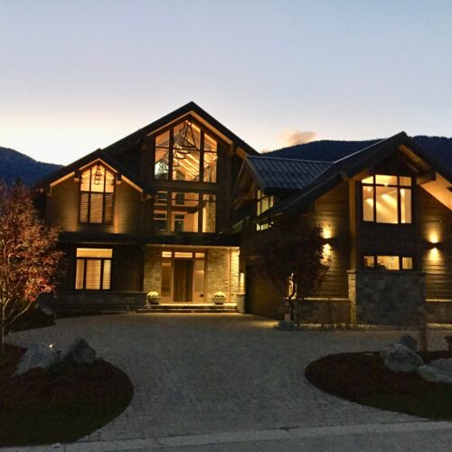 Modern two-story house with large windows and exterior lights on at dusk, surrounded by trees and mountains in the background.