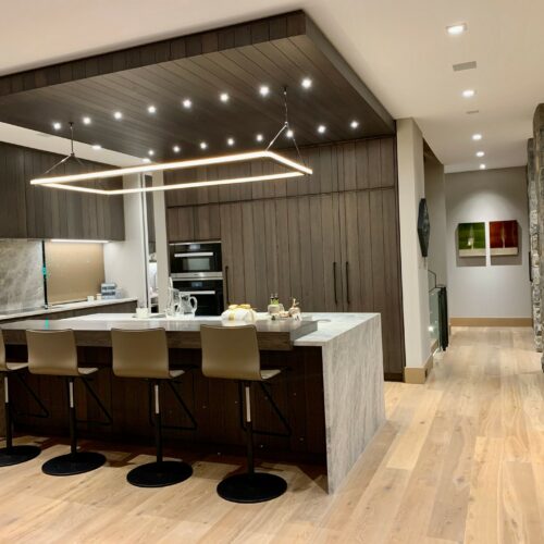 Modern kitchen with dark wood cabinets, a large island with four barstools, built-in ovens, a wine display wall, and pendant lighting above the island.