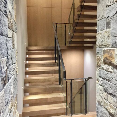 Modern wooden staircase with glass and metal railing, set between a stone wall and smooth light-colored walls.