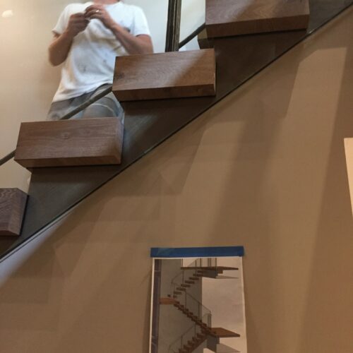 A person stands near a modern wooden staircase; below the steps, a taped architectural drawing of the staircase is attached to the wall.