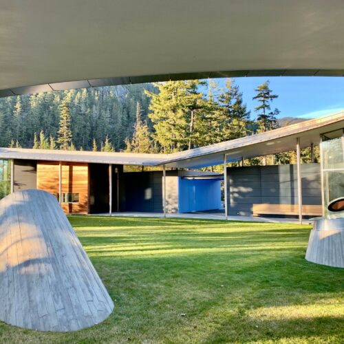 Modern house with curved rooflines, large glass windows, and abstract sculptures on a green lawn, set against a forested mountain backdrop.