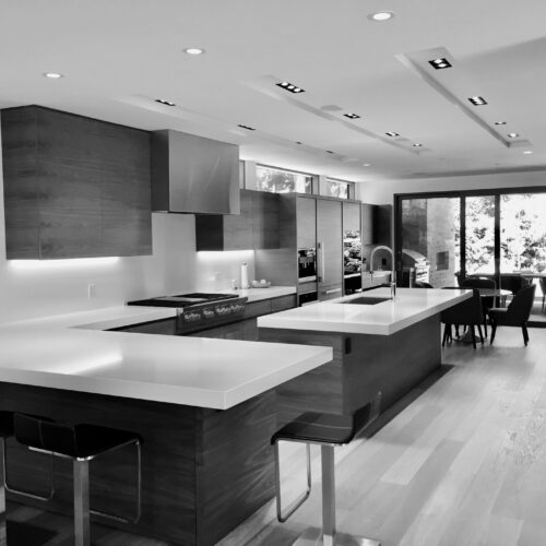 Modern kitchen with two large islands, bar stools, wood cabinetry, built-in appliances, and large glass doors opening to a dining area and outdoor view.