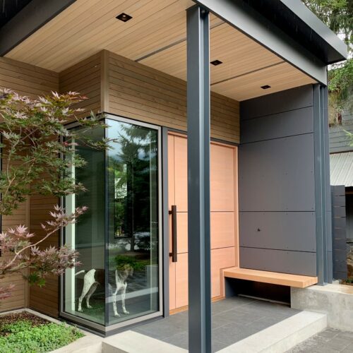 Modern house entrance with large glass windows, wood and metal finishes, a small bench, and a dog standing inside near the door.