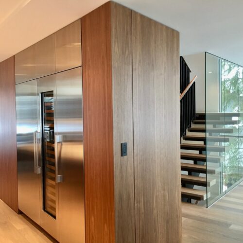 A modern interior features built-in stainless steel appliances in a wood-paneled wall next to an open staircase with glass railings.