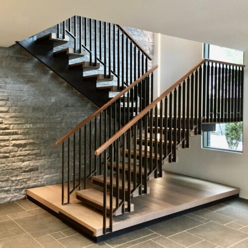 Modern indoor staircase with metal railings, wooden steps, and a stone accent wall, located in a well-lit space with large windows and tiled flooring.