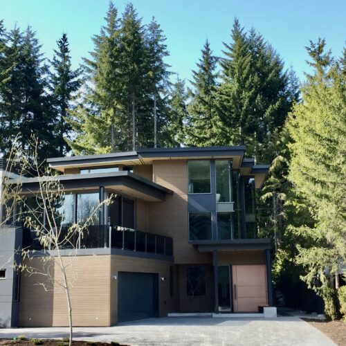 A modern three-story house with large windows and a flat roof, surrounded by tall evergreen trees under a clear blue sky.
