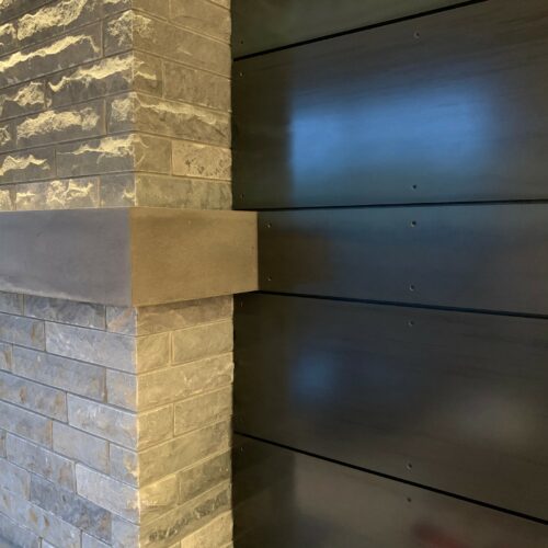 Close-up of a modern fireplace with gray stone bricks and a concrete mantel, next to a wall with dark horizontal wood panels.