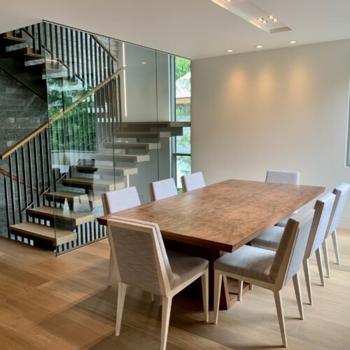A modern dining room with a wooden table surrounded by eight upholstered chairs, next to a floating staircase with glass railing and a large window.