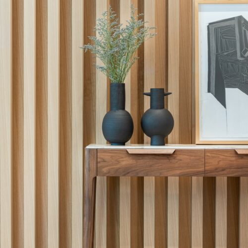 A wooden console table with two dark vases, a framed abstract artwork, and a plant, set against a vertical wooden slat wall.