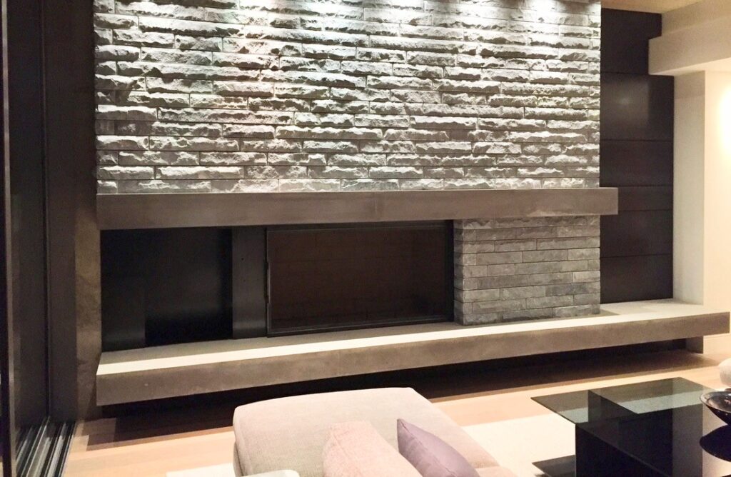Modern living room fireplace with a wide stone facade, horizontal concrete mantel, and dark metal accents, illuminated by recessed ceiling lights.