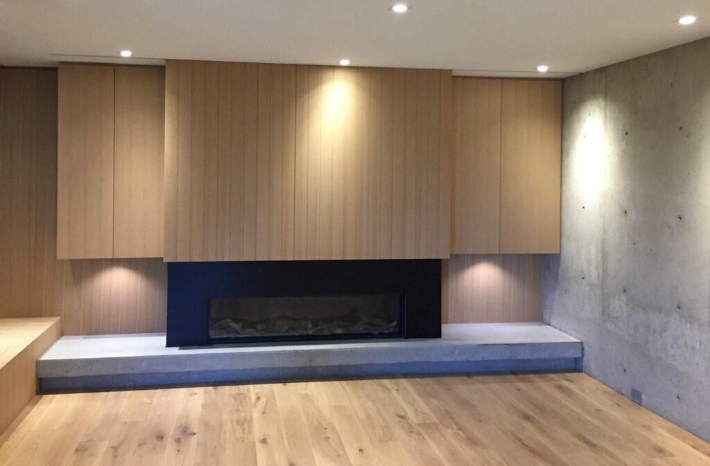 Modern living room with light wood wall panels, a black rectangular fireplace, concrete bench, and wood flooring under recessed ceiling lights.