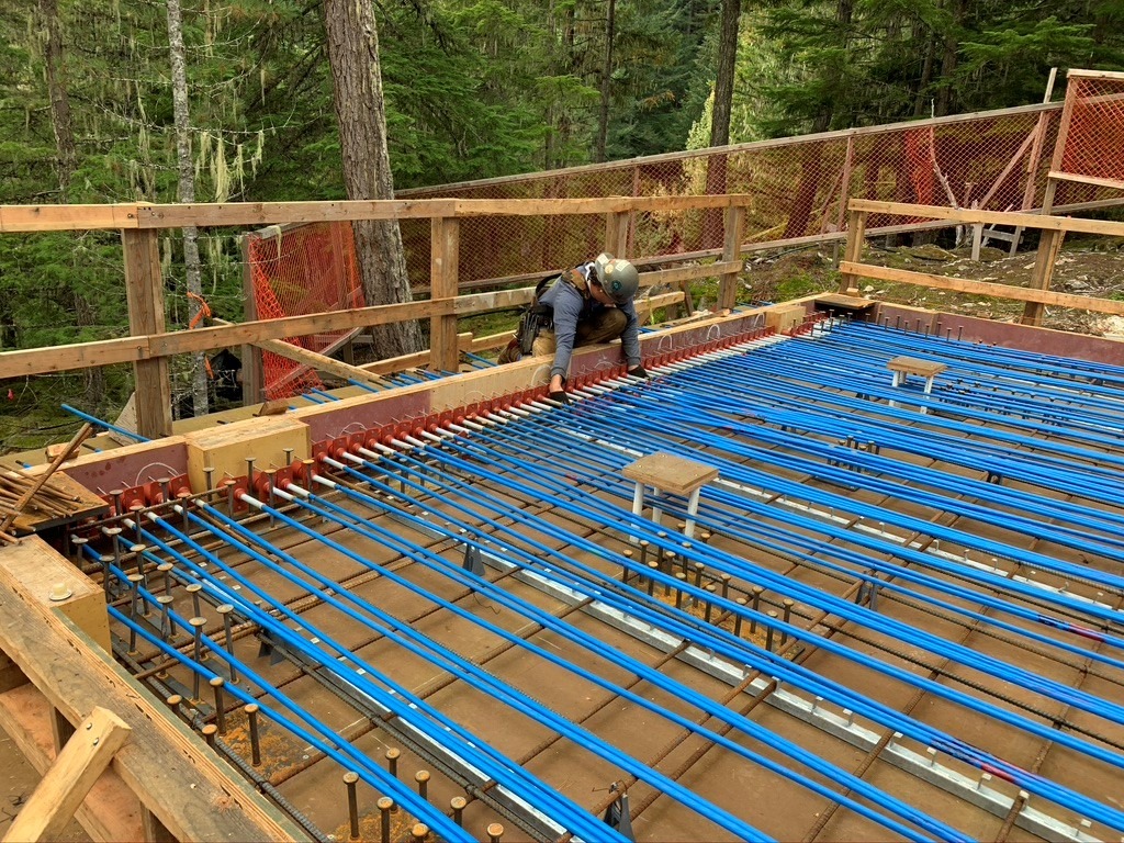 A construction worker installs blue rebar and other reinforcements on a wooden-framed concrete slab in a forested area.