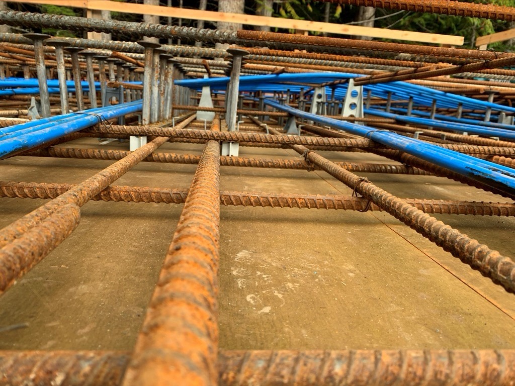 Close-up view of rusted steel rebar arranged in a grid pattern at a construction site, with blue pipes and wooden planks in the background.