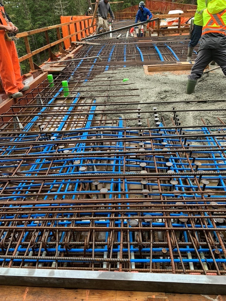 Workers standing on a bridge under construction with exposed rebar, blue conduit, and partially poured concrete. Construction materials and tools are visible.