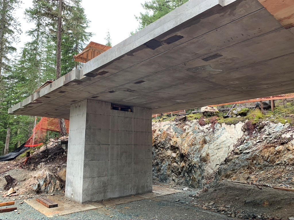 A concrete bridge under construction in a forested area, supported by a single central pillar, with exposed rock and trees in the background.
