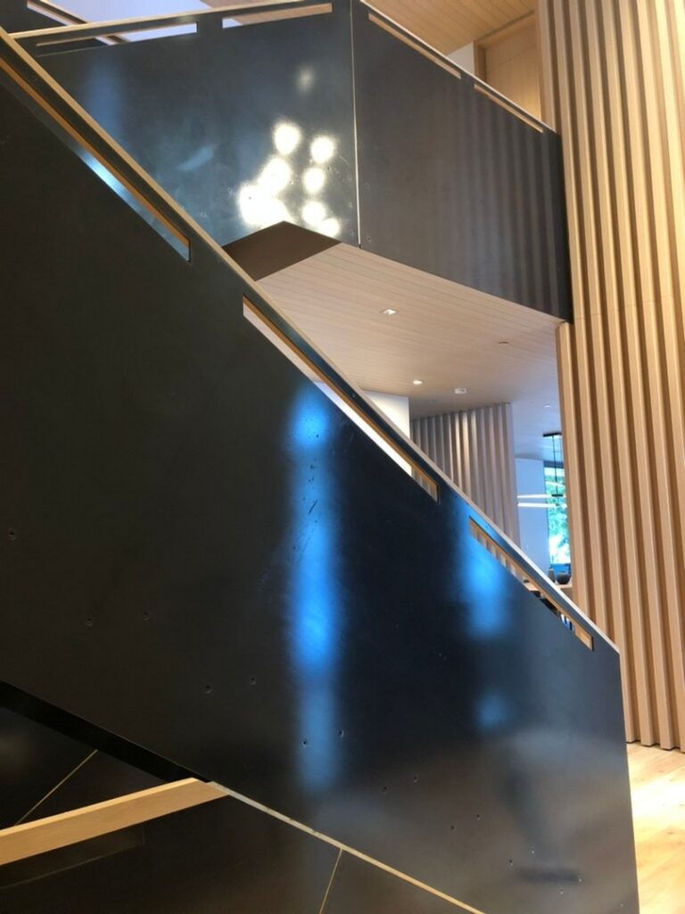 A modern interior staircase with black metal railings and wooden panel walls, illuminated by natural light and ceiling lights.